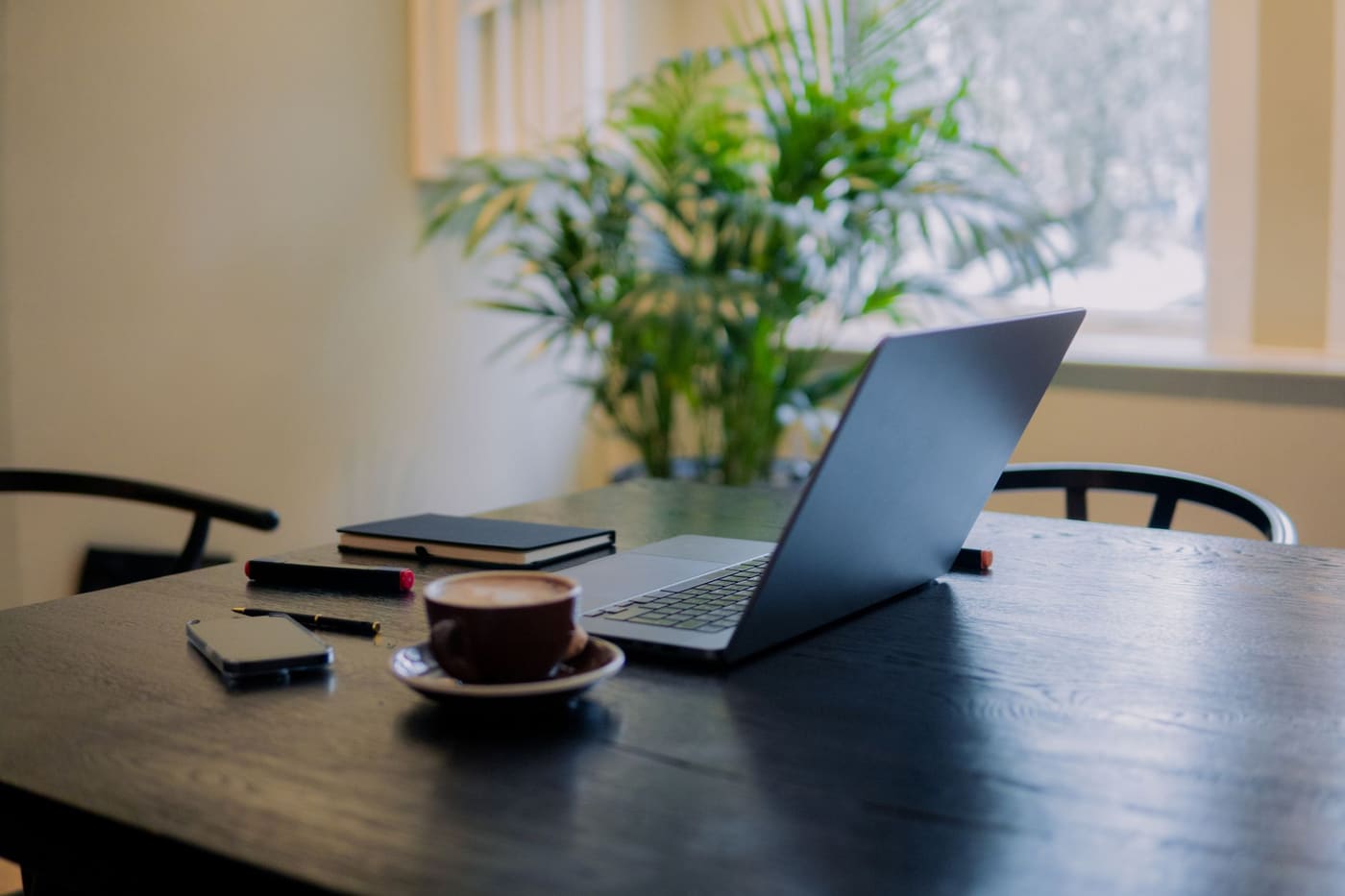 A laptop, coffee cup and phone on a dark wooden table by a window with a plant
